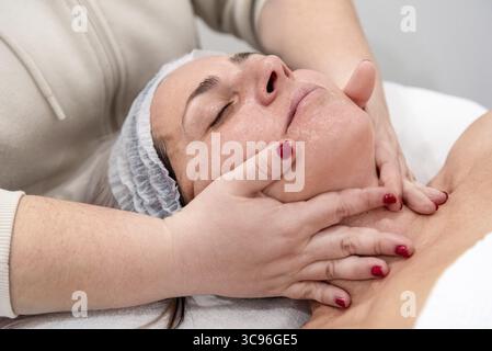 Estetista che applica crema idratante e massaggia viso e collo di una donna di mezza età sdraiata su una barella nel centro di bellezza Foto Stock
