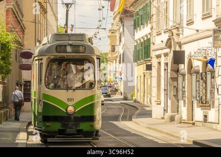 Vista di un tram d'epoca verde e crema scivola lungo le piste soleggiate, gettando lunghe ombre sui vecchi edifici, uno scorcio di un viaggio storico, Foto Stock
