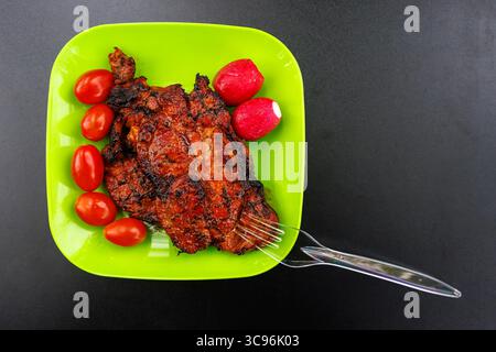 Piatto di plastica verde brillante con un pezzo di carne e verdure grigliate. Pomodori e ravanelli con carne. Piatto con carne su sfondo nero. Foto Stock