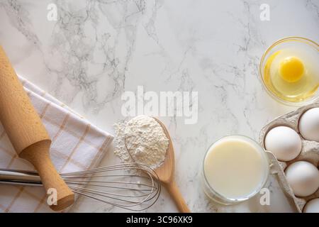 Farina, uova, latte e una frusta sono disposti ordinatamente su un bancone di marmo, preparandosi per una sessione di cottura in una cucina calda. Foto Stock