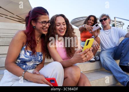 Gruppo di giovani che usano smartphone sulle scale, condividono contenuti divertenti e si divertono. Foto Stock