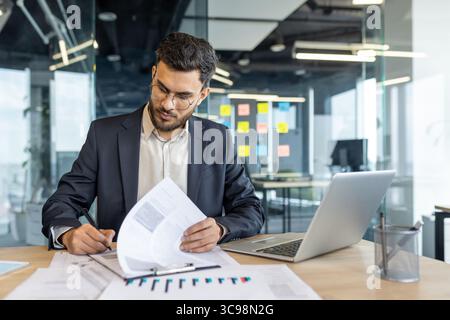 Un uomo d'affari lavora nel suo ufficio moderno, rivede documenti e utilizza il suo computer portatile. Concentrato e professionale, è vestito con un completo. Foto Stock