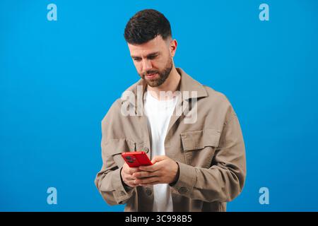 Il giovane barbuto scrive su uno smartphone rosso, indossa una giacca su sfondo blu Foto Stock