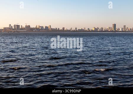 Lo skyline di Mumbai si estende attraverso l'orizzonte, visto dalle calme acque blu del mare sotto un cielo sereno, catturando la vivace costa urbana Foto Stock