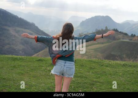 Vista posteriore di una donna avventurosa in piedi ai margini della scogliera di montagna, braccia aperte in condizioni di vento. Il clima nebuloso migliora l'atmosfera delle escursioni. Foto Stock