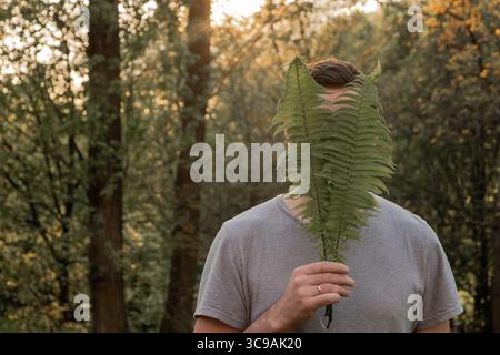Ritratto di un uomo caucasico senza volto in natura. Si copre la faccia con foglie di felce. Il concetto di unità con la natura e il benessere Foto Stock