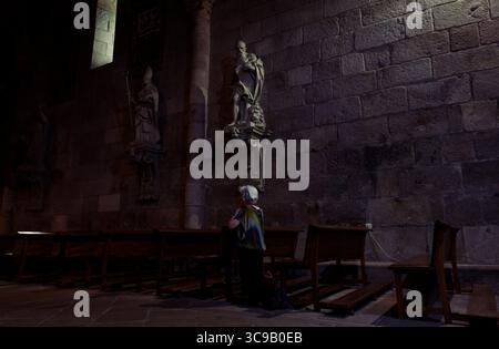 Donna inginocchiata in quieta preghiera sotto solenni santi in pietra in una chiesa poco illuminata a Braga Foto Stock