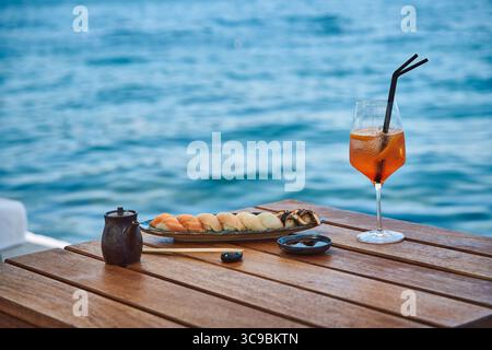 Deliziosa esperienza culinaria sull'acqua con sushi e cocktail rinfrescante su un tavolo di legno con una pittoresca vista sull'oceano Foto Stock