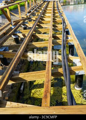 Vista dettagliata di un molo in legno in costruzione, che mostra travi, dispositivi di fissaggio, tubi e acqua poco profonda trasparente Foto Stock