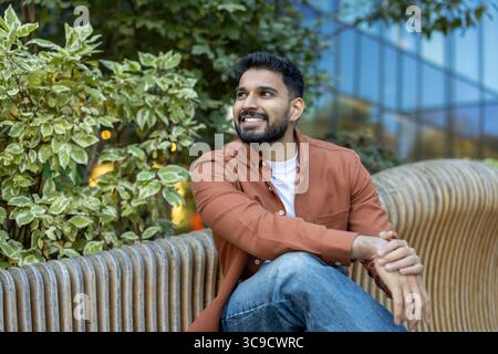 Un giovane e sorridente uomo siede all'aperto, godendosi una giornata di sole. Indossa un abbigliamento casual, circondato dal verde e dall'architettura moderna. Foto Stock