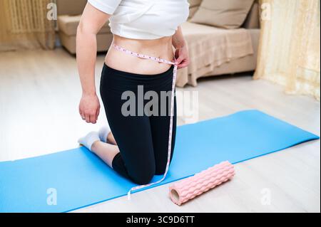 Donna con vita imperfetta che lo misura con il nastro mentre sta in piedi su un tappetino yoga, consapevolezza del corpo e concetto di cura personale Foto Stock