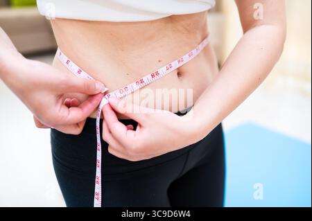 Donna con vita imperfetta che lo misura con il nastro mentre sta in piedi su un tappetino yoga, consapevolezza del corpo e concetto di cura personale Foto Stock