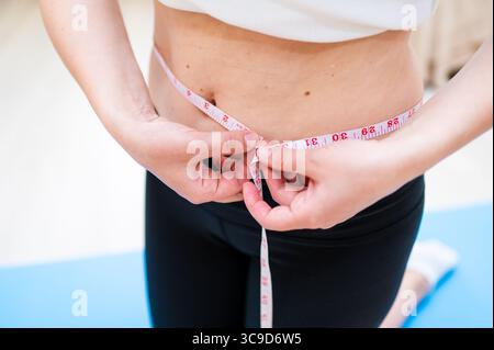 Donna con vita imperfetta che lo misura con il nastro mentre sta in piedi su un tappetino yoga, consapevolezza del corpo e concetto di cura personale Foto Stock