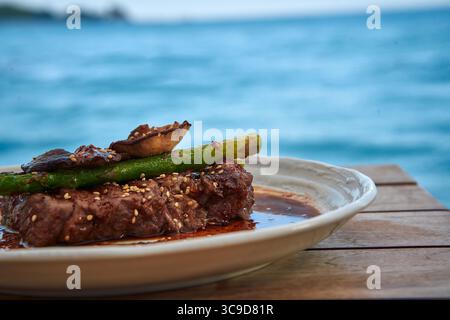 Un delizioso piatto di bistecca presentato ad arte con asparagi e funghi, sullo sfondo di una tranquilla vista sull'oceano, perfetto per gli amanti della cucina Foto Stock