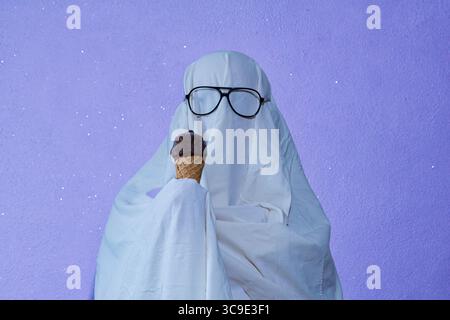 Un'immagine umoristica di una persona vestita con un semplice costume da fantasma fatto da un lenzuolo bianco, indossando occhiali da sole e tenendo in mano un cono gelato. Catturato Foto Stock