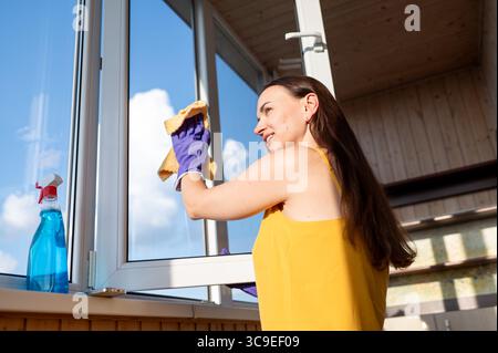Giovane casalinga felice che lava una finestra sotto la luce del sole, godendo delle faccende domestiche e dell'atmosfera fresca Foto Stock