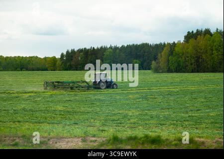 Trattore che taglia l'erba su un grande campo verde vicino a una foresta sotto un cielo nuvoloso, l'agricoltura in azione Foto Stock