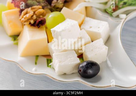 Piatto di formaggi del ristorante - vari tipi di formaggi con uva e olive nere su piatto bianco. Primo piano con messa a fuoco selettiva. Foto Stock