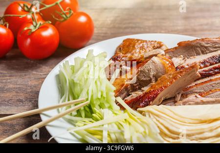 Carne di anatra cinese di Pechino servita con cipolle, pancake, pezzi di cetriolo e pomodori su piatto bianco con bacchette da vicino. Tavolo di legno. Foto Stock
