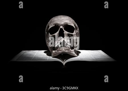 Cranio umano su un vecchio libro aperto su sfondo nero sotto un fascio di luce. Foto Stock