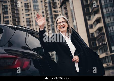 Donna d'affari sicura ed elegante sorridente mentre esce dall'auto sullo sfondo urbano durante il giorno Foto Stock