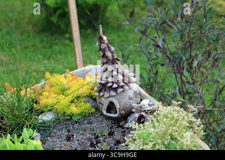 Una stravagante casa delle fate fatta di cono di pino si trova tra un sedum acro di pietra gialla, uno juncus effusus riccio e un fogliame verde in un lussureggiante piantagione Foto Stock
