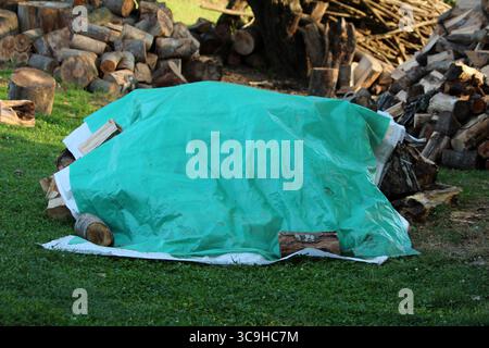 Una grande pila di legna da ardere tritata è protetta da un telo di plastica verde, circondato da tronchi e pezzi di legno su un prato erboso in un ambiente rurale Foto Stock