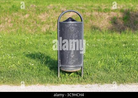 Un cestino in metallo perforato con coperchio a cupola si erge da solo su un'erba vibrante, promuovendo la pulizia in un ambiente urbano illuminato dal sole Foto Stock