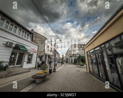 VALJEVO, SERBIA - 12 MAGGIO 2025: Via pedonale Kneza Milosa con negozi, caffetterie e residenti che camminano sotto cesti di fiori appesi e lampioni. Foto Stock
