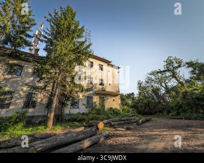 L'hotel abbandonato si trova in una radura forestale in Serbia. I tronchi caduti circondano l'edificio trascurato in cemento, con calda luce serale e alte conifere Foto Stock