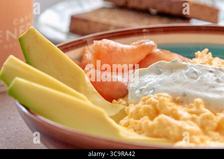 Una deliziosa ciotola per la colazione con uova strapazzate, salmone affumicato, avocado fresco e salsa di yogurt piccante servita con pane tostato Foto Stock