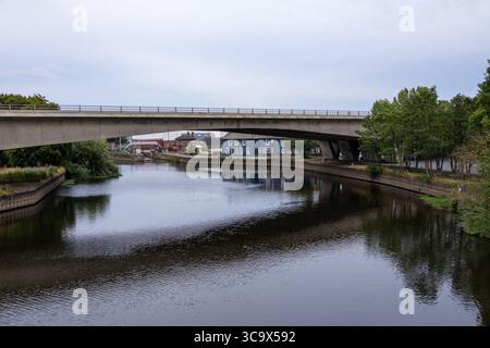 Moderno ponte di cemento che attraversa un tranquillo fiume riflettente, con alberi urbani e vegetazione lungo l'argine sotto un cielo nuvoloso. Foto Stock