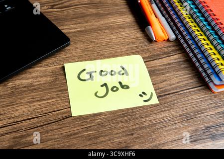 Nota adesiva con il messaggio "buon lavoro" e il volto sorridente sulla tastiera di un notebook. Quaderni colorati e penne su tavolo in legno. Foto Stock