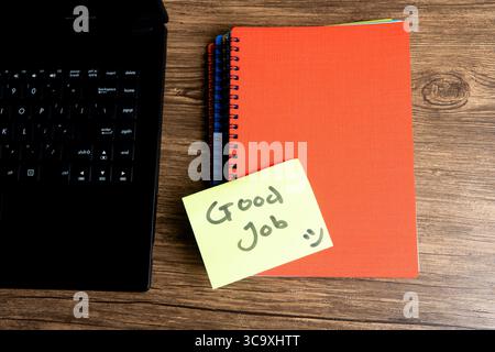 Nota adesiva gialla con "buon lavoro" e faccia sorridente su un notebook arancione. Tastiera per computer portatile visibile su una scrivania in legno. Foto Stock