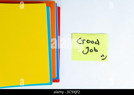 Primo piano di cartelle colorate e una nota adesiva gialla con il testo scritto a mano "Good Job" e la faccia sorridente su una parete bianca. Foto Stock