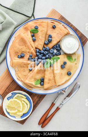 Frittelle sottili fatte in casa (crepes) con mirtilli freschi e panna acida su piatto bianco su sfondo chiaro, vista dall'alto Foto Stock