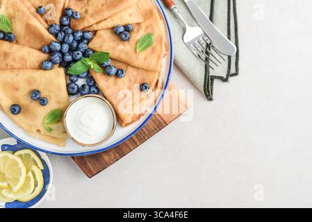 Frittelle sottili fatte in casa (crepes) con mirtilli freschi e panna acida su piatto bianco su sfondo chiaro, vista dall'alto Foto Stock