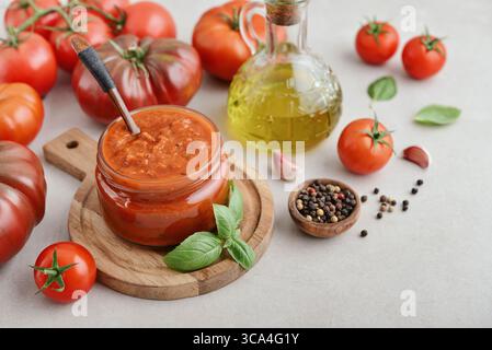 Salsa tradizionale di pomodoro in un vaso di vetro con basilico fresco, pomodori maturi, aglio e pepe su un tagliere di legno, primo piano Foto Stock