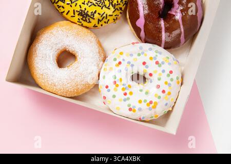 Primo piano di tre ciambelle colorate con glassa, sprinkles e zucchero a velo in una scatola di cartone su sfondo rosa pastello, varietà di ciambelle da caffè Foto Stock