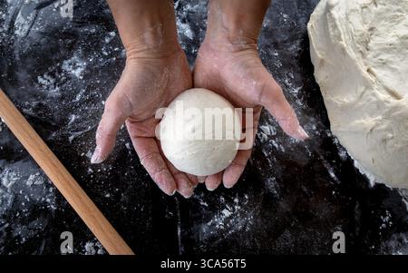Mani che tengono una palla di impasto fresco su un piano di lavoro nero infarinato. Preparazione fatta in casa di pane o pizza con farina e mattarello in vista. Cap Foto Stock