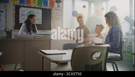 Madre e padre che guardano i risultati del test scolastico del loro figlio durante la serata dei genitori. Insegnante e genitori felici di eccellenti prestazioni accademiche e comportamento di Elementary School Boy. Movimento lento. Foto Stock