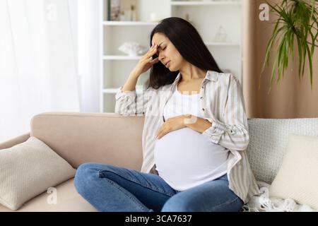Donna incinta che tocca la fronte con la mano, mostrando segni di mal di testa a casa Foto Stock