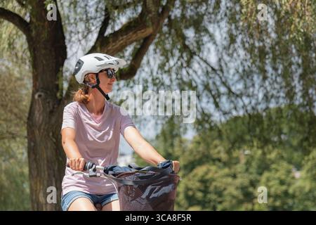 Una donna guida la sua bicicletta in una calda giornata sotto un albero, indossando un casco e occhiali da sole mentre si gode la vita all'aria aperta. Foto Stock