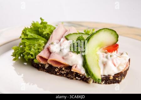 Classico smørrebrød danese con prosciutto salato, cetriolo croccante, lattuga fresca e condimento cremoso su una fetta di pane di segale scuro Foto Stock