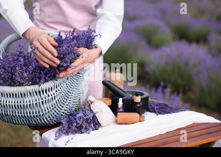 Fare cosmetici naturali. Donna con cesto in vimini di fiori di lavanda e altri ingredienti a tavola nel campo, primo piano. Spazio per il testo Foto Stock