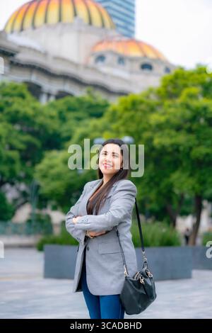 Giovane donna d'affari latina sorridente e felice che posa con le braccia incrociate di fronte al Palacio de Bellas Artes a città del Messico, con un blazer grigio e una pa blu Foto Stock
