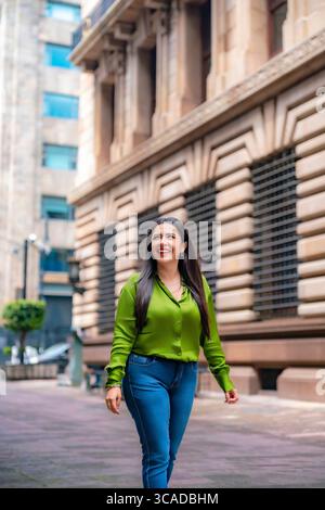Giovane e sicura donna latina che indossa una blusa verde brillante e jeans blu, cammina allegramente vicino al Banco de México nello storico centro finanziario Foto Stock