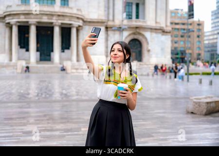La giovane latina sorridente con i capelli intrecciati, in una camicetta bianca ricamata e una gonna nera, scatta un selfie tenendo in mano un caffè nel centro storico di città del Messico. Foto Stock