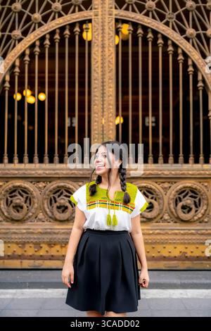 La giovane latina con i capelli intrecciati sorride brillantemente mentre si trova di fronte alle porte ornate del Palacio postale, indossando un tradizionale BL ricamato Foto Stock
