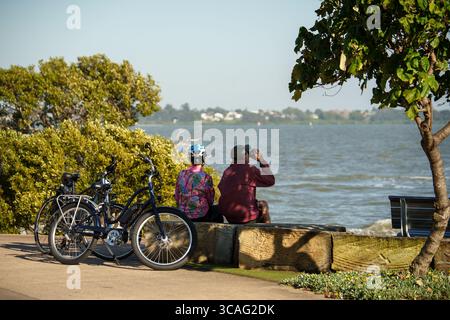 Due persone siedono sul lungomare con le biciclette parcheggiate nelle vicinanze, una con binocolo per godere della vista panoramica sulla costa in una giornata di sole. Foto Stock
